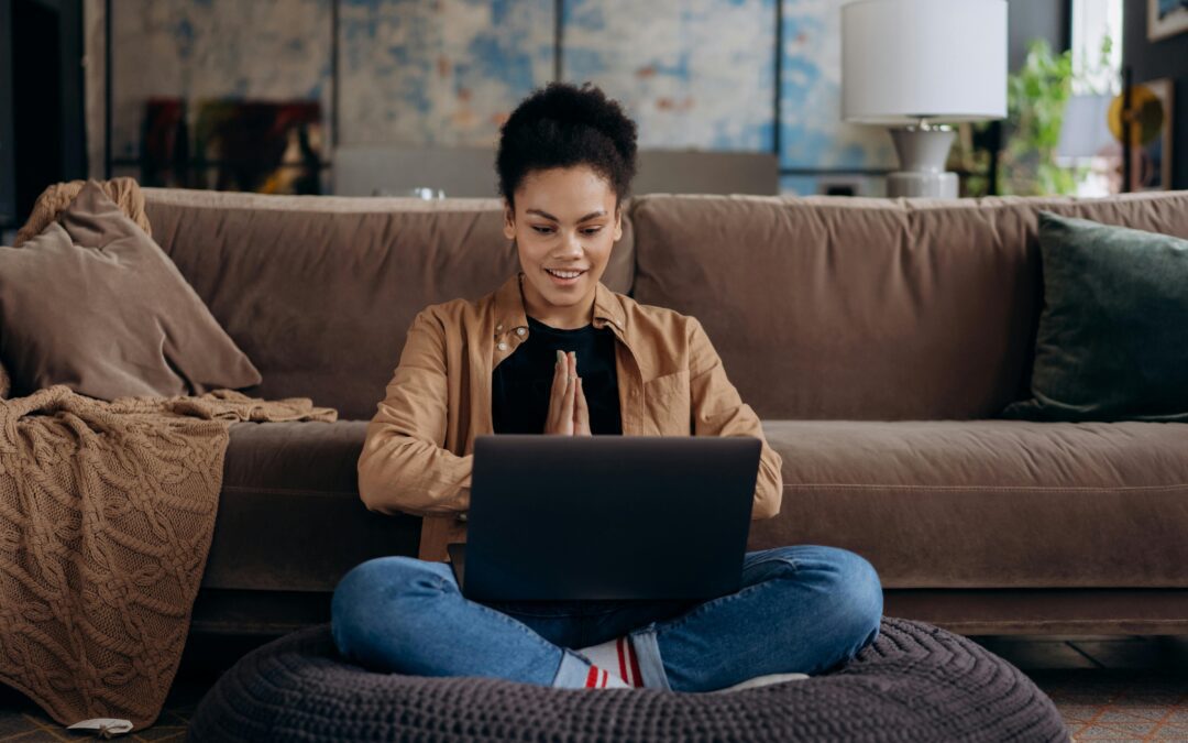 A smiling young woman sits cross-legged on a pouf, using her laptop in a cozy home setting.